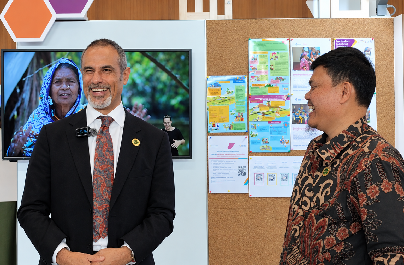 Two men stand side by side, both laughing. The man on the left wears a dark suit and paisley tie; the man on the right wears a brown batik shirt. Behind them, a screen shows a portrait of an older woman in a blue headscarf with a sign language interpreter in the corner, next to a corkboard of colourful posters on disability-inclusive disaster risk reduction.